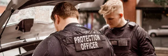 Two protection officers in tactical vests checking the open trunk of a vehicle in the rain